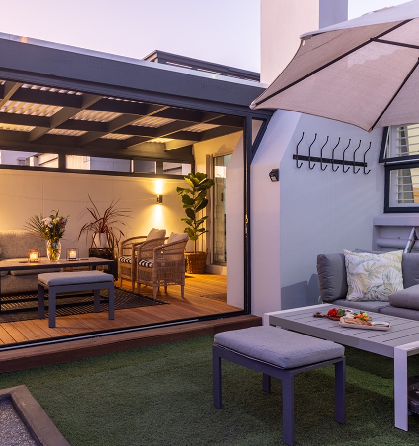 Covered patio at Atlantic Beach Cottage at dusk, with lounge seating, a parasol and warm lighting spilling from the open living room